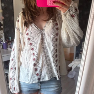 Vit blus med broderade blommor - Supersöt vit blus med broderade röda blommor och svarta detaljer. Långärmad med luftiga ärmar och v-ringning. Tunt, lätt material som känns perfekt för vår och sommar. Boho-vibe och snygg till både jeans och kjol.