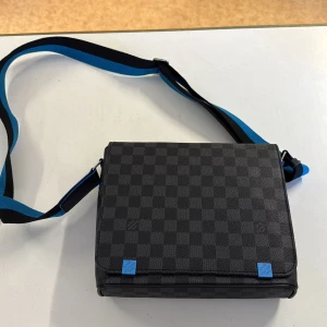 Louis Vuitton rutig axelväska svart/blå - Snygg axelväska från Louis Vuitton i svart och grått med klassiskt rutigt mönster. Väskan har en bred axelrem i svart och blått, samt blå detaljer. Insidan är fodrad i blått tyg och har innerfack. Perfekt för dig som vill ha en stilren och modern väska.