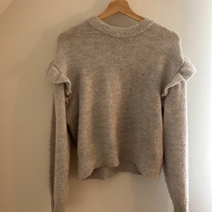 Beige stickad tröja med volang Gina Tricot - Supermjuk beige stickad tröja från Gina Tricot med volangdetaljer på axlarna. Tröjan har rund halsringning, ribbade muddar och lång ärm. Perfekt för dig som gillar en söt och enkel stil med en twist.