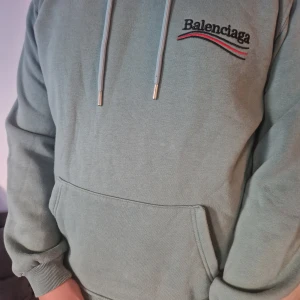 Balenciaga hoodie i grönt och svart - Två feta Balenciaga hoodies, en i dämpad grön med broderad logga och röd-svart detalj, samt en svart med stor vit logga på ryggen och mindre tryck fram. Båda har huva, snörning och känguruficka. Perfekt för dig som gillar streetstyle och vill sticka ut. 1800kr styck eller båda 3000kr