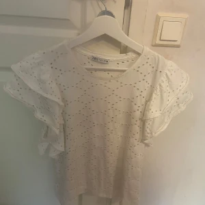 Vit blus med volangärm från Zara - Supersöt vit blus från Zara med broderade hål och volangärmar. Blusen har rund halsringning och är tillverkad i bomull, vilket gör den både luftig och bekväm. Perfekt för dig som gillar romantiska detaljer och en fräsch look.