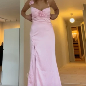 Ljusrosa långklänning med rosdetalj - Superfin ljusrosa långklänning i satinliknande material med smala axelband och snygg slits framtill. Klänningen har en söt rosdetalj vid bysten och korsade band i ryggen. Perfekt för bal eller student! Sitter figurnära och ger en elegant siluett.