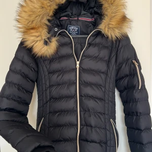 Svart dunjacka med fuskpäls från Vinson - Snygg svart dunjacka från Vinson Polo Club med fluffig fuskpäls på huvan. Jackan har guldiga dragkedjor och fickor med dragkedja. Perfekt för kalla dagar och ger en riktigt clean look.
