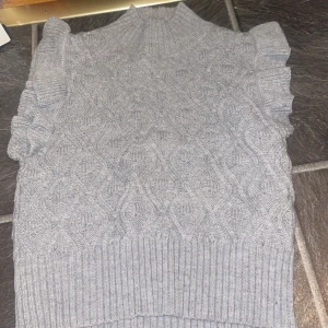 Grå stickad ärmlös tröja med volang - Supersöt grå stickad tröja med hög hals och volangdetaljer vid axlarna. Tröjan är ärmlös och har ett snyggt stickat mönster över hela plagget. Perfekt att styla med skjorta under eller som den är för en trendig look.