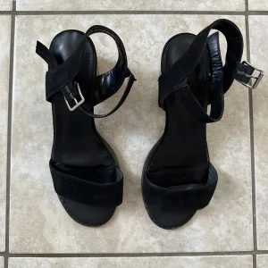 Svarta vår sommar sandaler sandeltter klackar - Stilrena svarta sandaler med bred rem över tårna och justerbart spänne runt ankeln. Skorna har en stabil, låg träfärgad klack och ovandel i mocka. Perfekta för dig som vill ha en enkel men snygg sko med lite höjd.