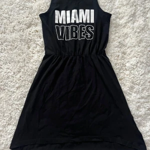 Svart klänning Miami Vibes KappAhl - Svart ärmlös kortklänning från KappAhl med coolt tryck 'MIAMI VIBES' i vitt på bröstet. Klänningen har resår i midjan för en snygg passform och är gjord i mjuk bomull. Perfekt för en chill sommardag eller när du vill ha en avslappnad look.