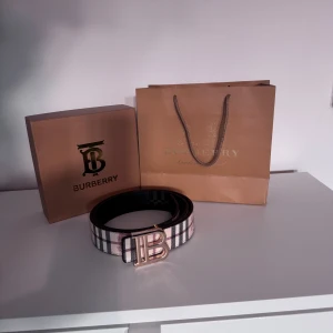 Burberry rutigt bälte med B-spänne - Snyggt Burberry bälte med klassiskt rutigt mönster i beige, svart och vinrött. Bältet har ett stort metallspänne i form av ett B och är gjort i skinn. Perfekt accessoar för att lyfta din outfit med en lyxig touch.