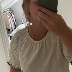 Vit blus med svarta detaljer från Zara - Superfin vit blus från Zara i storlek 164. Blusen har korta puffärmar med svarta broderade detaljer vid ärmslut och smockade partier på axlarna. Materialet är lätt och krinklat, vilket ger en avslappnad och trendig look.