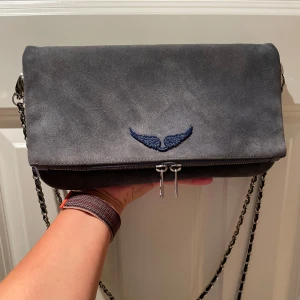 Grå mocka axelväska Zadig & Voltaire - Snygg blå/grå axelväska i mocka från Zadig & Voltaire med silverfärgad kedjerem och blått  vingemblem på framsidan. Väskan är i mycket gott skick, kan skicka fler bilder privat om det önskas. 