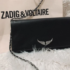 Zadig & Voltaire svart axelväska - Snygg svart axelväska från Zadig & Voltaire i skinn med silverfärgad kedjerem och ikonisk vingdetalj framtill. Aldrig använd