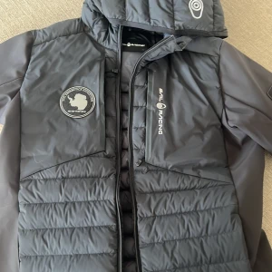 Svart Sail Racing dunjacka med huva - Snygg svart dunjacka från Sail Racing med broderad patch på bröstet och logga på bröst och huva. Jackan har quiltad framsida, dragkedja och huva. Perfekt för kyliga dagar och har en sportig vibe.