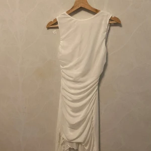 Hell ny Vit långklänning med drapering - Säljer hell ny, Snygg vit långklänning med draperad effekt baktill. Klänningen är ärmlös och har en elegant, enkel design som ger en stilren look. Perfekt för dig som vill sticka ut med en klassisk och tidlös stil. Storlek M