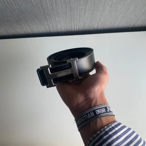 Svart bälte skärp - Säljer ett stilrent blått skärp med ikoniskt H-spänne i silverfärgad metall. Bältet har vita kontrastsömmar och är gjort i skinn. Perfekt accessoar för att lyfta din outfit med en lyxig touch.