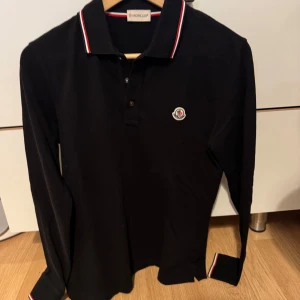 Svart Moncler pikétröja i ull - Svart långärmad pikétröja från Moncler i 100% ull. Klassisk krage med röd och vit randig detalj samt matchande detaljer vid ärmslut. Broderad Moncler-logga på bröstet. Perfekt för dig som gillar stilrena och exklusiva plagg.