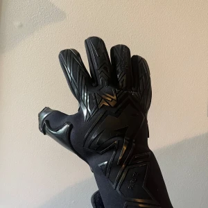 Svarta målvaktshandskar från RINAT - Säljer ett par svarta målvaktshandskar från RINAT med coolt blixtmönster på ovansidan och greppig insida. Handskarna har lång mudd med kardborre och är tillverkade i syntetmaterial. Perfekta för dig som vill sticka ut på fotbollsplanen. Helt nya!! Storlek 10 och pris 500kr