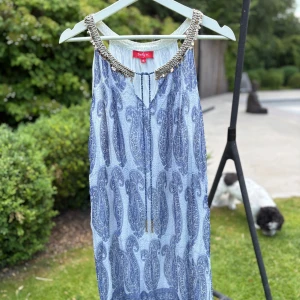 Blå paisleymönstrad klänning Derhy - Supersnygg blå och vit kortklänning från Derhy med paisleymönster, dekorativt halsband med detaljer och knyt framtill. Ärmlös modell i luftig bomull, perfekt för varma dagar. Ger en bohemisk och chill vibe. Liten i storleken!