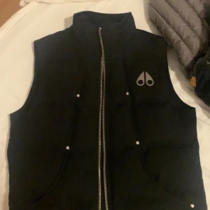 Svart dunväst Moose Knuckles - Svart dunväst från Moose Knuckles med hög krage och silverfärgad dragkedja. Västen har två fickor med tryckknappar och ikonisk logga i metall på bröstet. Insidan har broderad detalj och innerficka med dragkedja. Perfekt för lager-på-lager-stil.