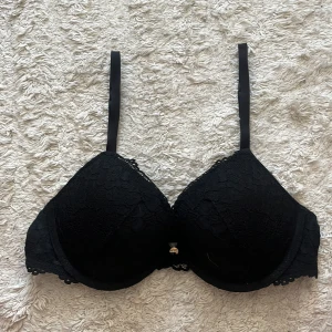 Svart spets-bh från H&M 75B - Svart bh från H&M i storlek 75B med snygg spetsdetalj över kuporna och tunna, justerbara axelband. Bh:n har en liten guldfärgad detalj framtill och klassisk knäppning bak. Perfekt för dig som gillar en stilren och feminin look.