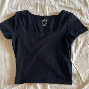 Svart croppad topp från Hollister - En svart, squareneck croppad topp från Hollister med fyrkantig ringning och korta ärmar. Enkel och stilren design i mjuk bomull, perfekt att matcha med jeans eller kjol. Snygg passform som framhäver axlarna och ger en modern look.