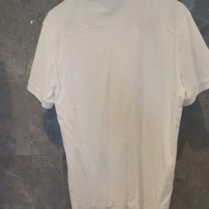 Vit pikétröja från J.Lindeberg - Klassisk vit pikétröja från J.Lindeberg med korta ärmar och krage. Tröjan har knappar framtill och diskret JL-logga på bröstet. Tillverkad i bomull för en skön och luftig känsla. Perfekt för en clean och stilren look.