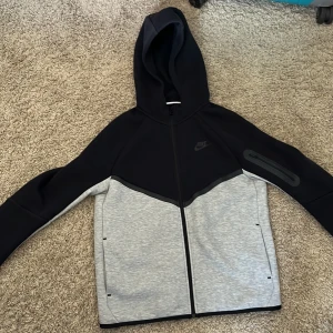 Nike tech svart grå XL 158-170 - Nike hoodiejacka i svart och ljusgrått med hel dragkedja och huva. Jackan har en sportig look, praktisk ficka med dragkedja på ärmen och diskret Nike-logga på bröstet. Perfekt för chill dagar eller när du vill ha en clean streetstyle.