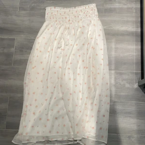 Vit blommig långkjol från Gina Tricot - Superfin vit långkjol från Gina Tricot med små rosa blommor och smockad midja. Kjolen har knytband med tofsar och är gjord i ett lätt, luftigt material. Stolen 170 och säljs inte längre!