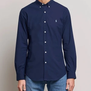 Mörkblå skjorta från Ralph Lauren - Klassisk mörkblå skjorta från Ralph Lauren med button-down krage och vita knappar. Skjortan har lång ärm och den ikoniska broderade loggan på bröstet. Perfekt till jeans eller chinos för en clean och stilren look. Tappat lite färg