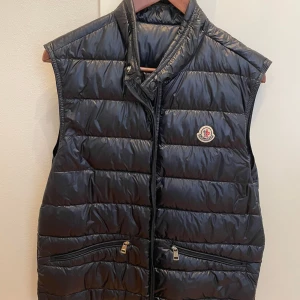 ÄKTA Moncler Gui väst - En riktig fin och elegant väst som passar till både sommaren och hösten!                Västen är i princip i nyskick förutom att en bit av dragkedjan har gått av som kan se på en av bilderna. Men har dock sparat den vilket betyder att den går att återställa till nyskick!                                                           Både kvitto och alla tags som man fick med från NK finns kvar också! Kostar idag ca 9000 att köpa från butik. Storlek 3 motsvarar ungefär storlek M.