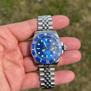 Seiko blå dykarklocka i stål - Snygg Seiko dykarklocka med blå urtavla och vridbar blå bezel. Automatisk rörelse och datumfönster med förstoringsglas. Robust länkarmband i rostfritt stål och tydliga indexmarkeringar. Vattentät upp till 300 meter, perfekt för dig som gillar stilren design och funktion.