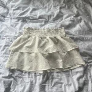 Vit volangkjol med smock - Supersöt vit kortkjol med smock i midjan och dubbla volanger. Kjolen har en luftig känsla och är perfekt för varma dagar. Den är enkel att matcha och ger en fräsch look.