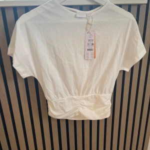 Vit croppad topp från Gina Tricot - Säljer en fräsch vit croppad topp från Gina Tricot. Toppen har korta ärmar, rund halsringning och en snygg rynkad detalj i midjan som ger en cool passform. Perfekt till jeans eller kjol för en clean look.