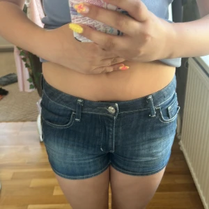 Mörkblå jeansshorts med fickor - Snygga mörkblå jeansshorts med klassisk femficksdesign och låg midja. Shortsen har silverfärgade knappar och dragkedja framtill samt coola sömdetaljer. Perfekta för sommardagar och lätta att matcha med allt.