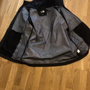 Svart vindjacka från The North Face - Säljer en svart vindjacka från The North Face med DryVent-teknologi. Jackan har hel dragkedja, meshfoder och logga på bröstet och ryggen. Perfekt för blåsiga dagar och enkel att slänga på sig över en hoodie. Klassisk stil med elastiska ärmslut.