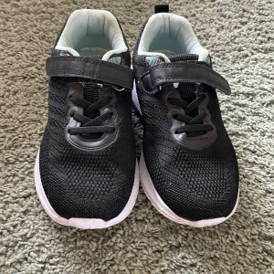 Svarta sneakers med kardborreband - Snygga svarta sneakers med vit sula och praktiskt kardborreband. Ovandelen är i mesh-liknande syntetmaterial som andas och har elastiska snören för enkel påtagning. Insidan är ljusblå för en fräsch kontrast. Perfekta för en aktiv vardag eller gympaskor!