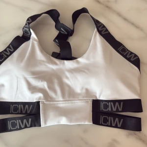 Vit sportig topp från ICANIWILL - Snygg vit sporttopp från ICANIWILL med svarta elastiska band och logga runt axlar och midja. Toppen är ärmlös och har en modern, atletisk look som passar perfekt till träning. Materialet är mjukt och följsamt för maximal komfort.
