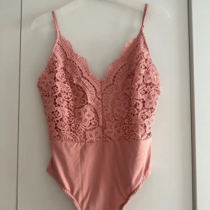 Rosa spetsig body med blommönster - Supersöt rosa bodysuit med tunna axelband och djup v-ringning. Överdelen är täckt av detaljerad spets med blommönster och underdelen är i mjuk bomull. Perfekt för dig som vill ha en feminin och trendig look med extra detaljer