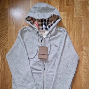 Grå hoodie från Burberry med dragkedja - Snygg ljusgrå hoodie från Burberry med klassiskt rutigt foder i huvan och insidan. Tröjan har dragkedja framtill, snörning i huvan och broderad logga på bröstet. Perfekt för dig som gillar stilrena och exklusiva plagg med en twist.