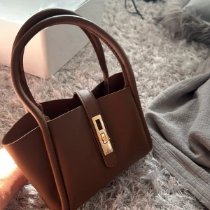 Brun handväska med gulddetalj - Snygg brun handväska i skinnimitation med två rejäla handtag och guldfärgat spänne framtill. Väskan har stilren design och är perfekt för att bära dina viktigaste grejer. Enkel att matcha med olika outfits och har en klassisk look. Axelband kommer med. 