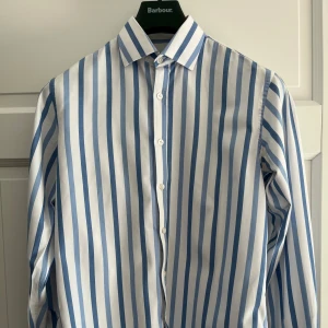 Blåvit randig skjorta La Chemiserie - Snygg skjorta från La Chemiserie Traditionnelle med breda blå och vita ränder. Klassisk krage, knäppning framtill och lång ärm. Perfekt för dig som gillar en stilren och fräsch look. Tillverkad i bomull för en skön känsla hela dagen. Intialer på ena manschetten som enkelt går att ta bort. 