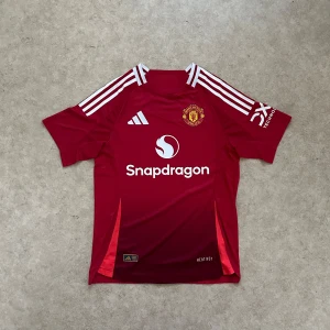 Manchester United hemmatröja 2024/25 - Röd Manchester United fotbollströja från Adidas med vita detaljer och logga på bröstet. Kortärmad modell med klassiska tre ränder på axlarna och sponsortryck på bröst och ärm. Tillverkad i lätt och ventilerande funktionsmaterial.