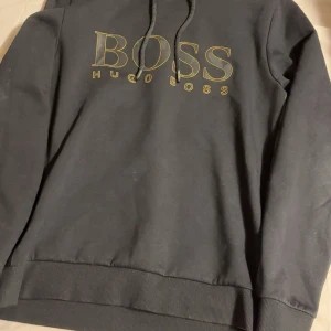 Svart hoodie från Hugo Boss - Svart hoodie från Hugo Boss med stort BOSS-tryck i guld på bröstet. Tröjan har huva med snörning och ribbade muddar. Snygg detalj med svart mönstrad rand längs ärmen. Perfekt för en chill och stilren look.