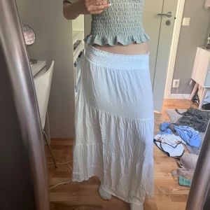 Vit långkjol med smock i midjan - Superskön vit långkjol med bred smock i midjan och lätt rynkad nederdel. Kjolen har en flowy och luftig känsla, perfekt för varma dagar. Passar dig som gillar en avslappnad och bohemisk stil.