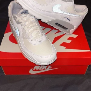 Nike Air Max 90 vita sneakers - Nike Air Max 90 sneakers i vitt med grå detaljer och klassisk siluett. Skorna har en synlig Air Max-enhet i sulan, snörning och reflekterande Swoosh-logga på sidan. Ovandelen är i syntet och textil för en clean look. Perfekta för dig som gillar stilrena och sportiga vibes. Aldrig använt dem helt rena och orörda 