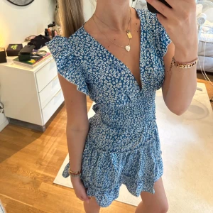 Blå blommig kortklänning med volang - Supersöt blå kortklänning med vitt blommönster, volangärmar och v-ringning. Klänningen har öppen rygg med knyt och volangdetaljer i kjolen. Den har två små hål vid sömmen på kjolen. Lätt att fixa med antingen sy eller bara sätta en säkerhetsnål har funkat jättebra 