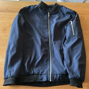 Mörkblå bomberjacka med dragkedja - Snygg mörkblå bomberjacka med klassisk siluett, ribbade muddar och silverfärgad dragkedja framtill. Aldrig använd. Liten L