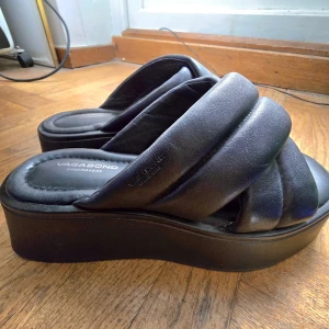 Svarta platåsandaler från Vagabond - Chunky svarta sandaler från Vagabond med breda, vadderade remmar i skinn och hög platåsula. Snyggt korslagda band och öppen tå ger en modern vibe. Perfekta för dig som gillar statement-skor och vill ha något bekvämt men ändå trendigt.