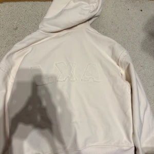 Beige hoodie från LXA med broderi - Säljer en stilren beige hoodie från LXA med broderade bokstäver på ryggen. Tröjan har huva, långa ärmar och är gjord i mjuk bomull. Perfekt för dig som gillar en clean och enkel look.