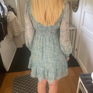 Blommig ljusblå kortklänning - Supersöt ljusblå kortklänning med blommigt mönster och v-ringning. Klänningen har långa, transparenta ärmar och volangkant nertill. Har endast använt denna klänning en gång så den är i nyskick💗Pris kan diskuteras.