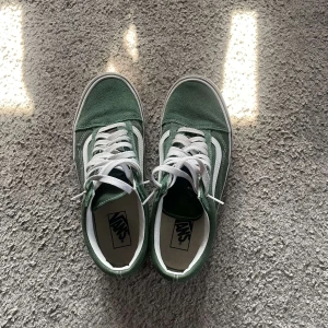 Gröna Vans sneakers - Säljer ett par klassiska Vans sneakers i grön mocka med vita detaljer och vit sula. Skorna har den ikoniska sidoremsan, vita snören och den bruna gummisulan med rutmönster. Perfekta för en chill streetstyle-look. Ända tecknet på användning är sulan på skon, som blivit missfärgad! Box medföljer.