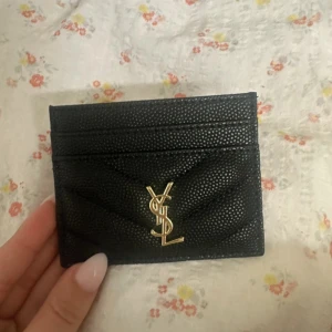 Svart YSL plånbok i skinn - Säljer en svart plånbok från YSL med guldigt logotyp framtill. Plånboken är i skinn med quiltad struktur och har flera kortfack. Perfekt storlek för att ha med sig överallt och riktigt snygg detalj i väskan.